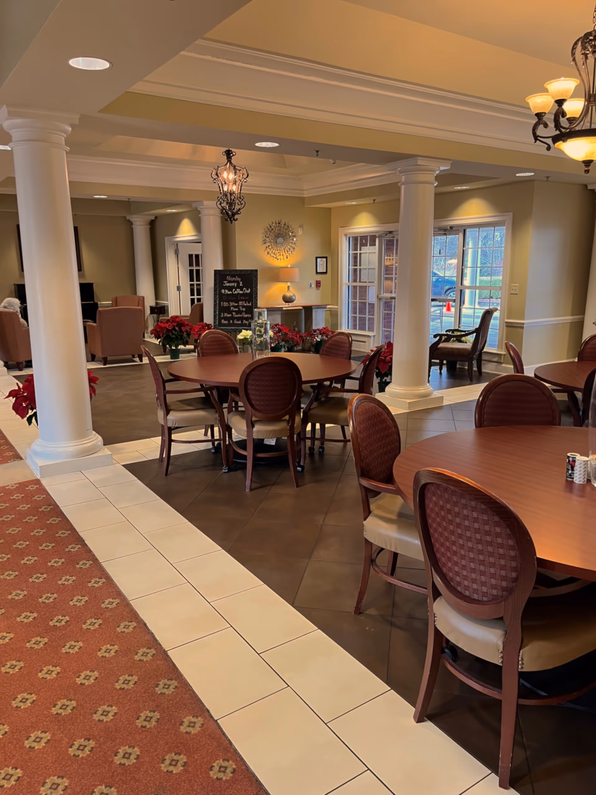 A spacious dining area in an assisted living facility with round wooden tables and cushioned chairs. The room features large white columns, warm lighting from chandeliers, and windows letting in natural light. There are red poinsettia plants placed around the room, and a small chalkboard with a schedule is visible in the background.