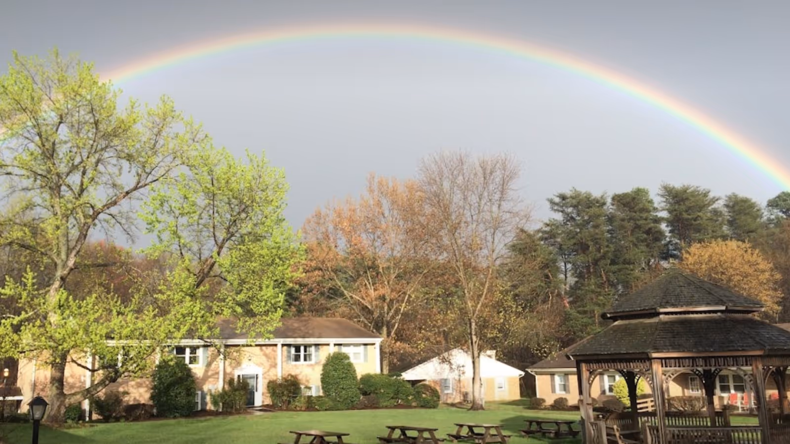 A rainbow arches over the Bello Machre outdoor campus, with trees, lawn, and a gazebo.