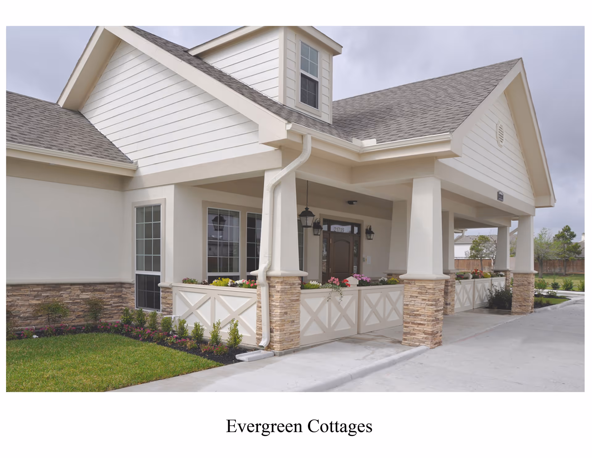 A beige senior living building with a covered front porch, stone pillars, and flowering planters.