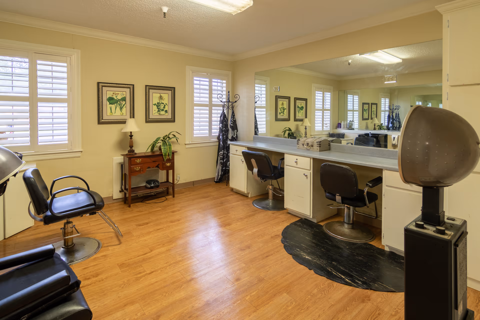 Interior view of a salon area in a skilled nursing facility with wooden flooring, two black salon chairs in front of a long counter with a large mirror, a hair dryer chair, a small wooden table with a lamp and plant, and framed botanical pictures on the wall.