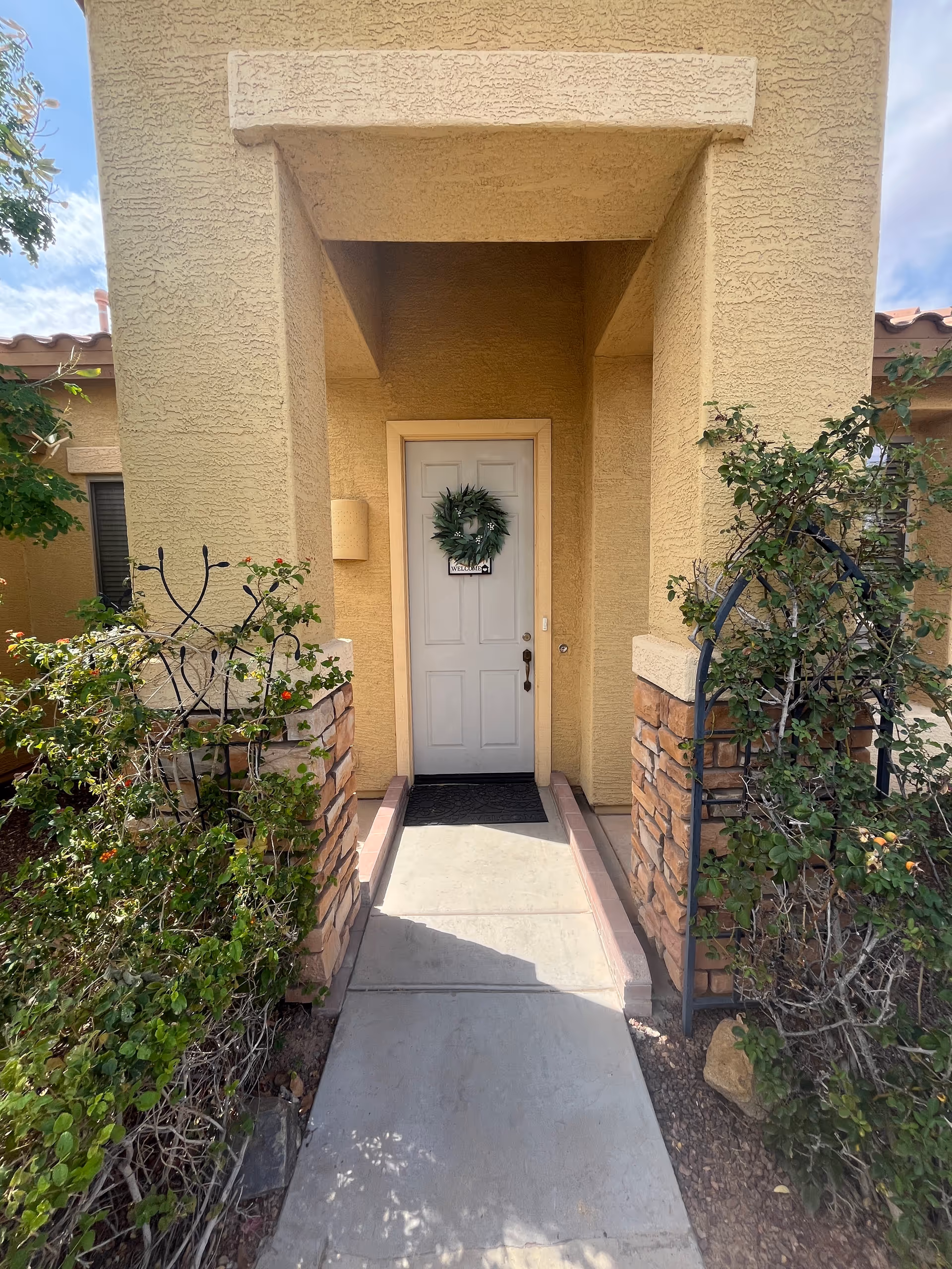 Beige stucco entrance with white door and wreath, flanked by plants and stone accents.