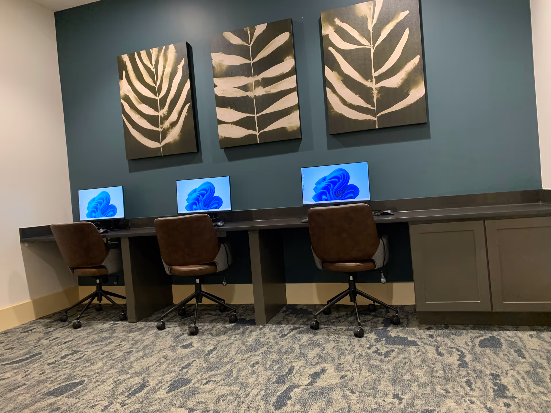 Row of computers on a long desk beneath teal wall with leaf artworks.