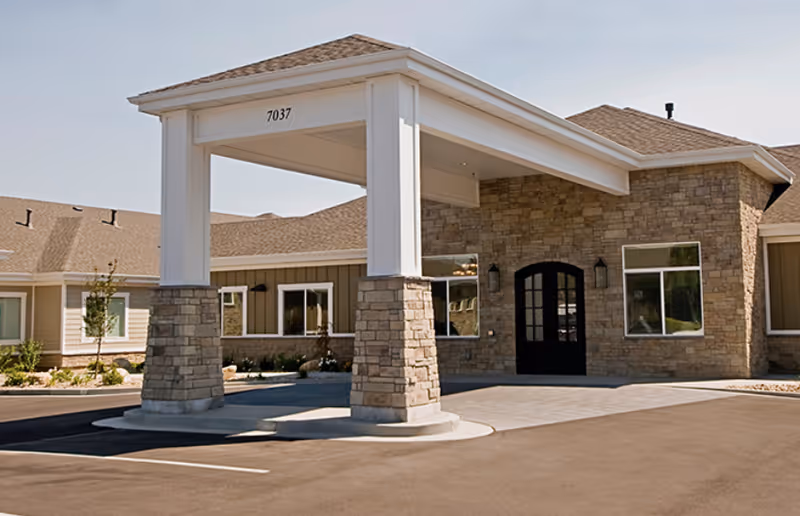 Front entrance of a single-story assisted living building with a stone facade and a covered porte-cochere displaying the number 7037.