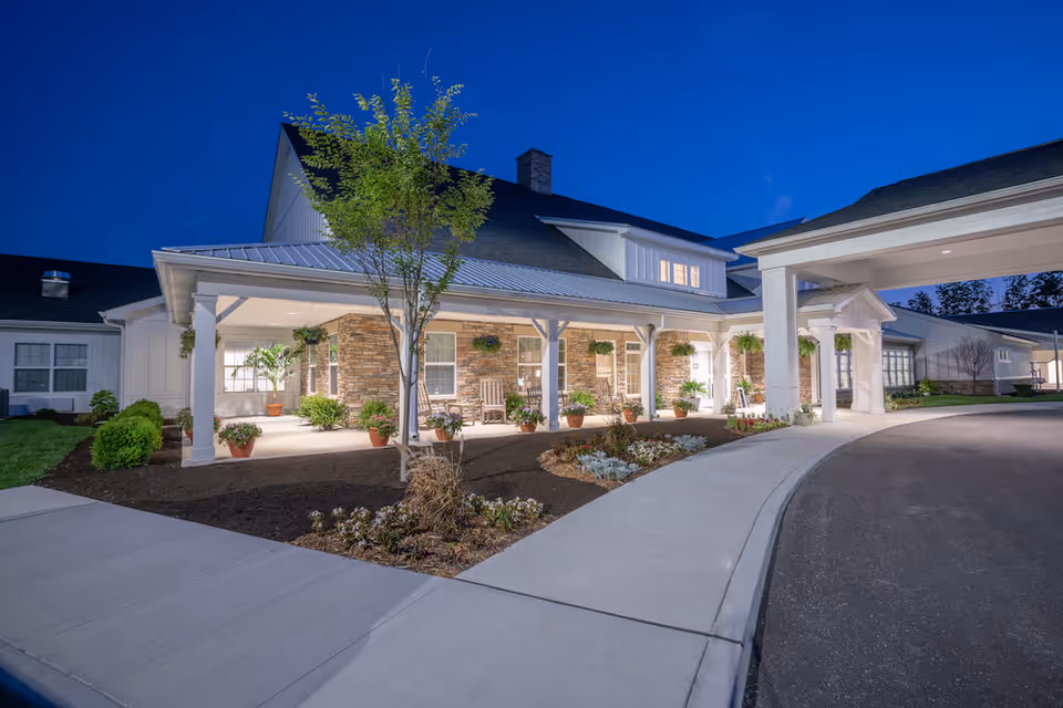 Covered, well-lit front entrance and porch of a senior living building at dusk with potted plants and a curved driveway.