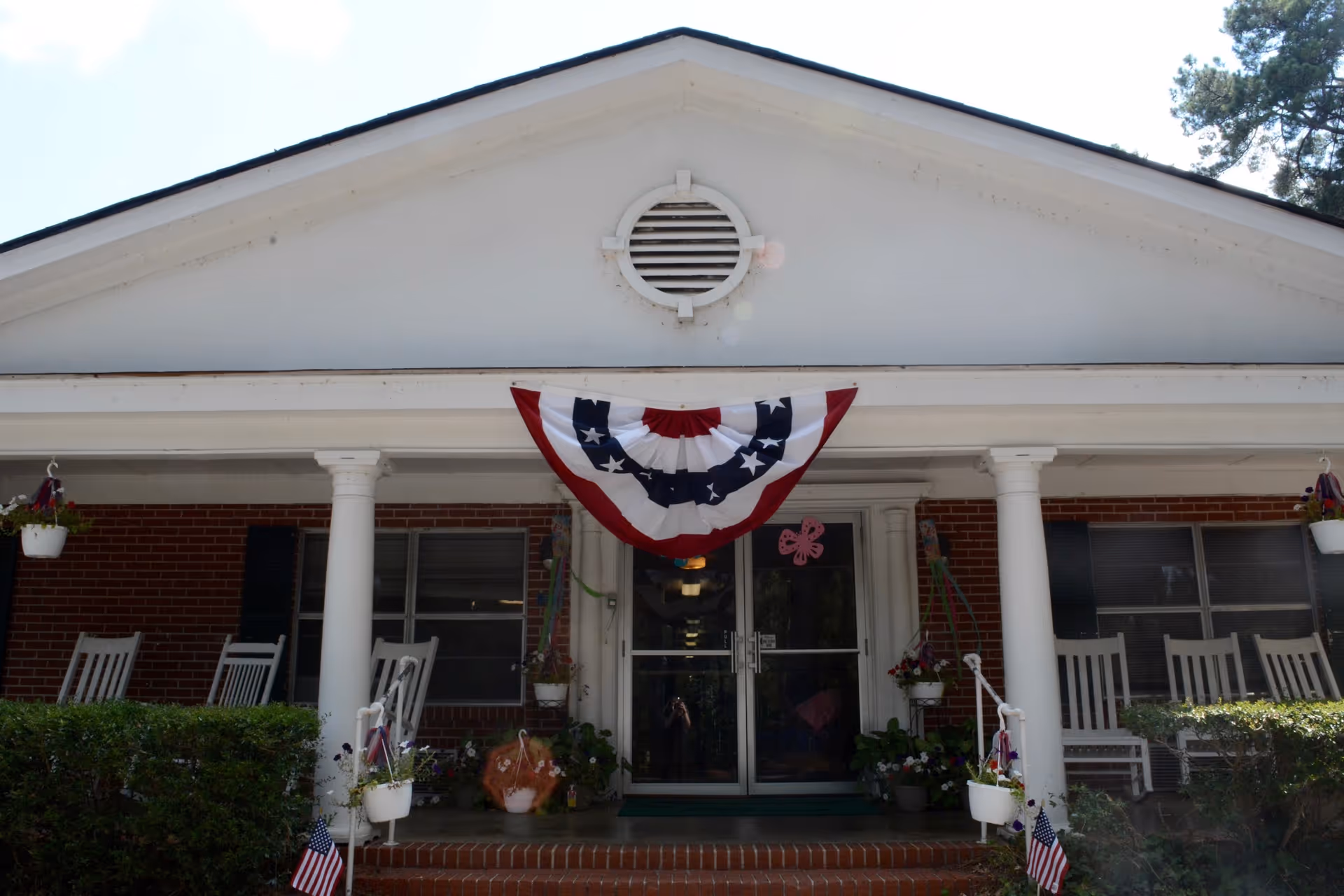 Front entrance of a nursing and rehabilitation facility with a white triangular pediment, two white columns, and a patriotic red, white, and blue bunting hanging above the double glass doors. There are white rocking chairs on the porch and potted plants hanging and placed around the entrance.