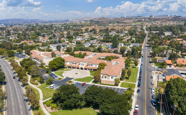 Aerial view of a senior living facility named Bayshire Yorba Linda surrounded by trees and residential neighborhoods. The facility has a large, U-shaped building with a circular driveway and parking areas. Roads with parked cars and houses extend into the distance under a partly cloudy sky.