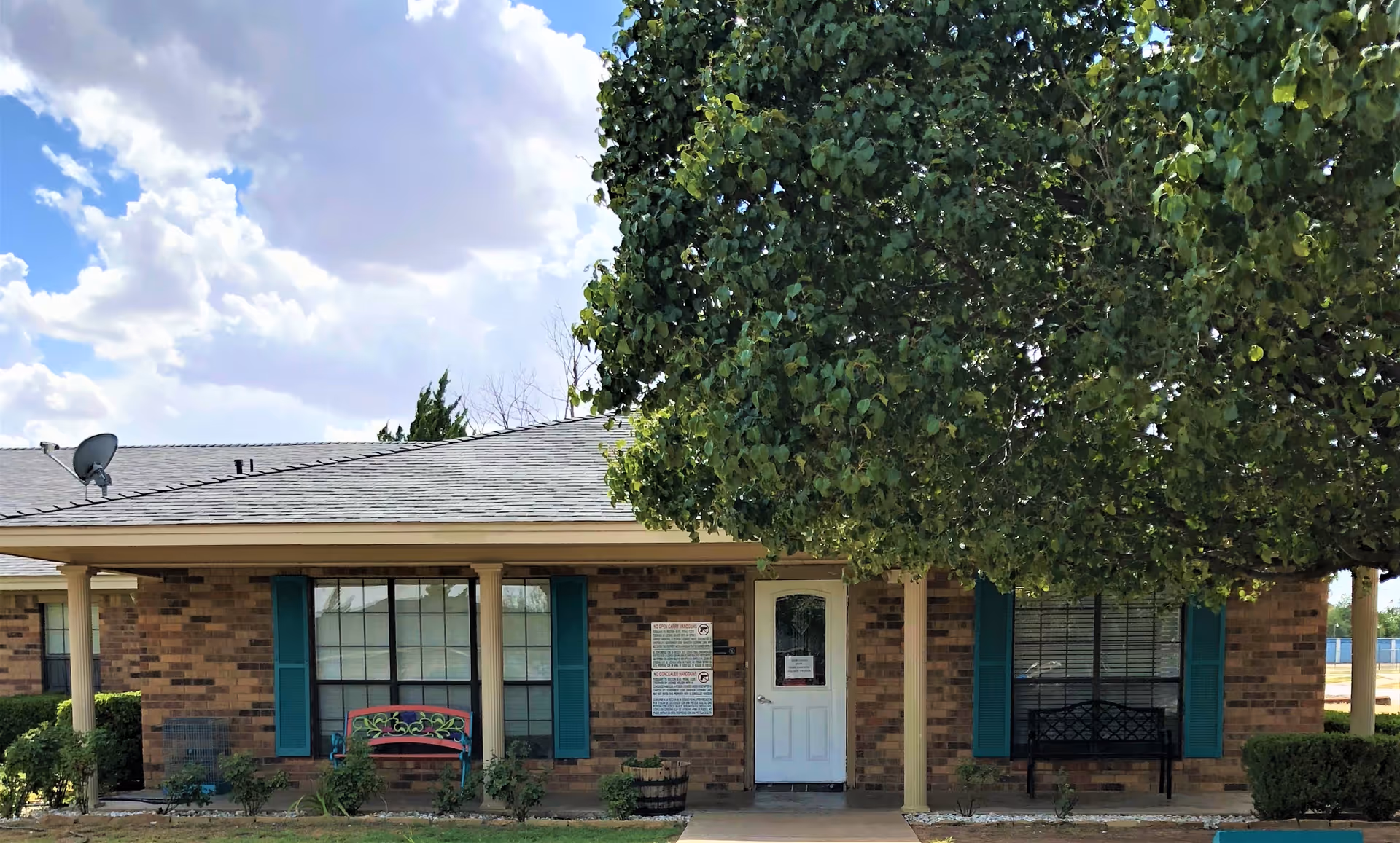 Front exterior of a single-story brick senior living building with a covered porch, white entry door, teal shutters, benches, and a large tree.