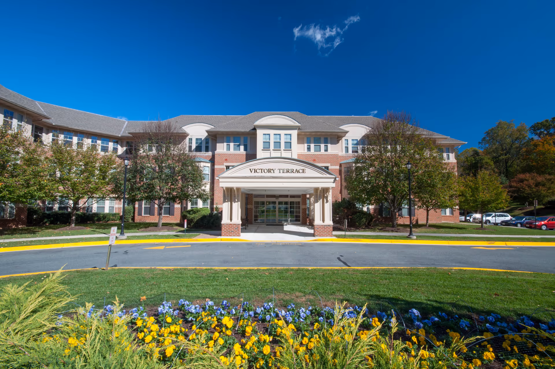 Front exterior view of Victory Terrace senior living facility with a covered entrance, brick and beige facade, surrounded by trees and a flower bed with yellow and purple flowers in the foreground under a clear blue sky.