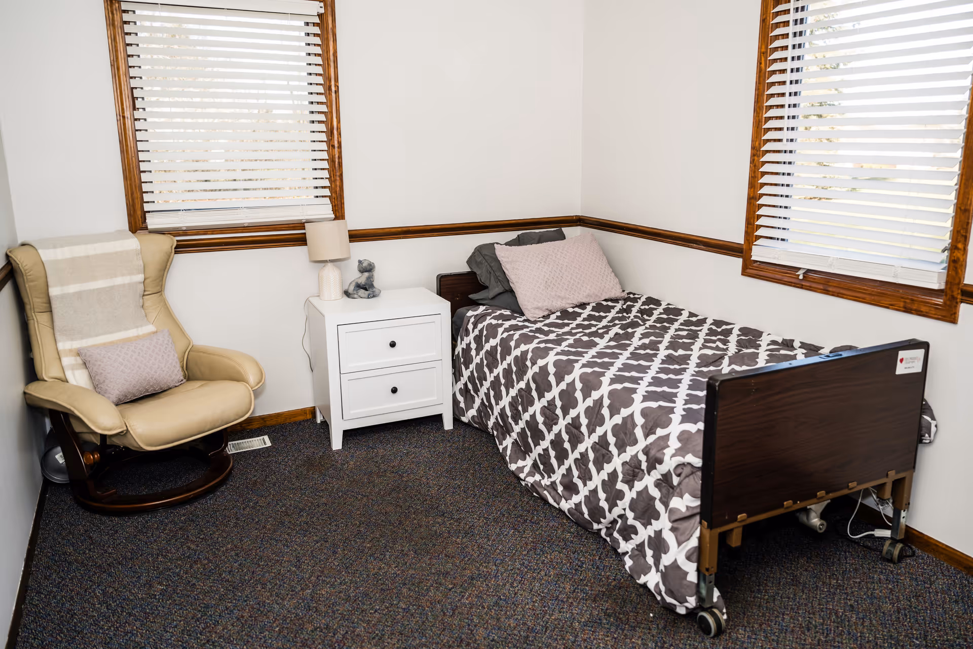 A small bedroom with a single hospital-style bed covered with a patterned gray and white comforter and two pillows. Next to the bed is a white nightstand with two drawers, a lamp, and a small decorative item. To the left is a beige cushioned armchair with a striped throw blanket and a small pillow. The room has two windows with white blinds and wooden trim, and the floor is carpeted.