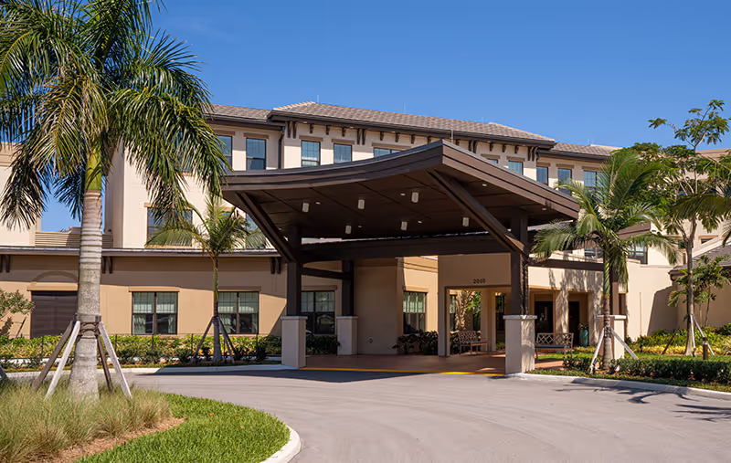Front entrance with a large covered porte-cochère of a multi-story building surrounded by palm trees and a driveway.