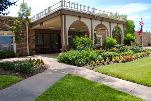 Entrance to a brick healthcare building with an arched covered walkway, landscaped lawn, and American and Texas flags.
