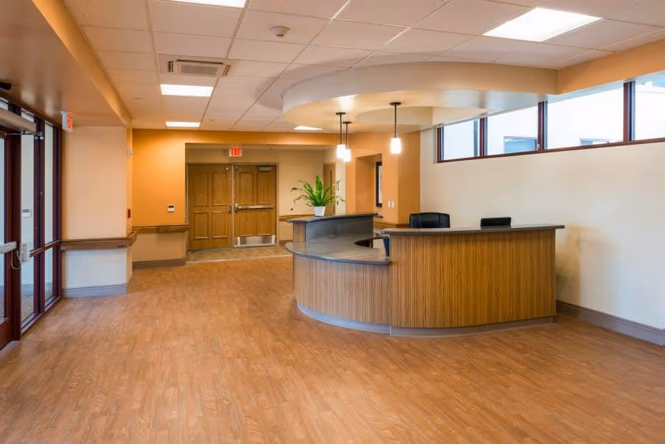 Spacious senior living facility lobby with curved wooden reception desk, plant, and warm lighting.