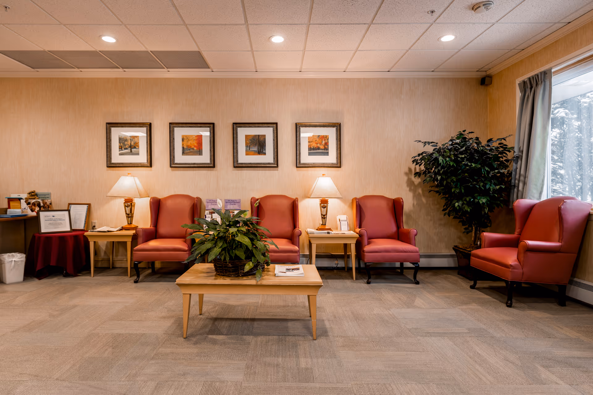A warmly lit senior living lobby with red chairs, plants, and framed art.