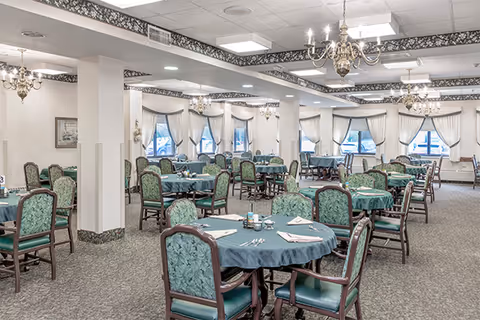 A spacious senior dining room with round tables, teal chairs, chandeliers, and draped windows.