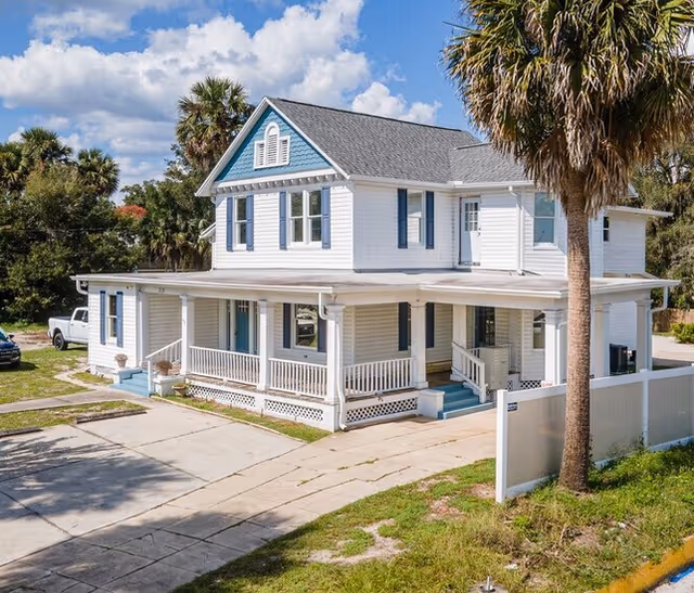 Two-story light-blue senior living home with wraparound porch and palm trees outside.