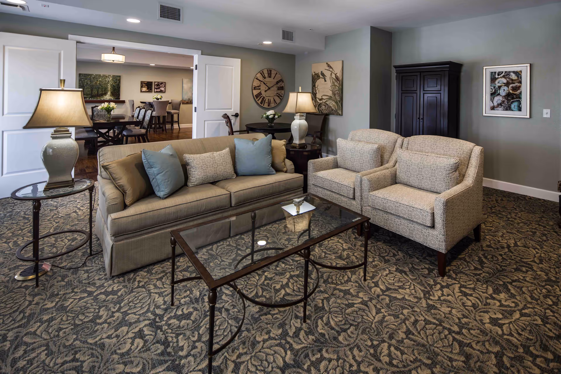 A furnished lounge with a sofa, two armchairs, a glass coffee table and lamps, opening to a dining area through double doors.