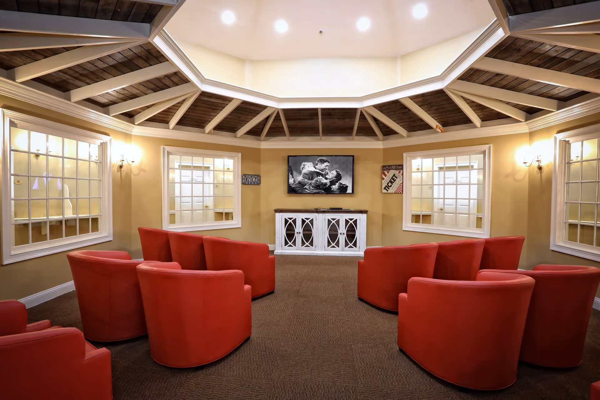 A cozy senior care facility media room with eight red armchairs arranged in two rows facing a flat-screen TV mounted on a beige wall. The ceiling features a wooden octagonal design with white beams and recessed lighting. There are four large windows with white frames on the walls, and two vintage-style movie posters on either side of the TV. The floor is carpeted in brown.