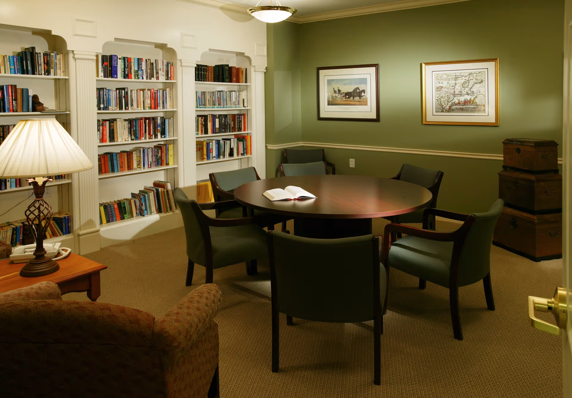 A cozy reading or meeting room with a round wooden table surrounded by six green upholstered chairs. There is an open book on the table. The room has built-in white bookshelves filled with books, a table lamp on a side table, two framed pictures on the green walls, and a stack of three wooden trunks in the corner.
