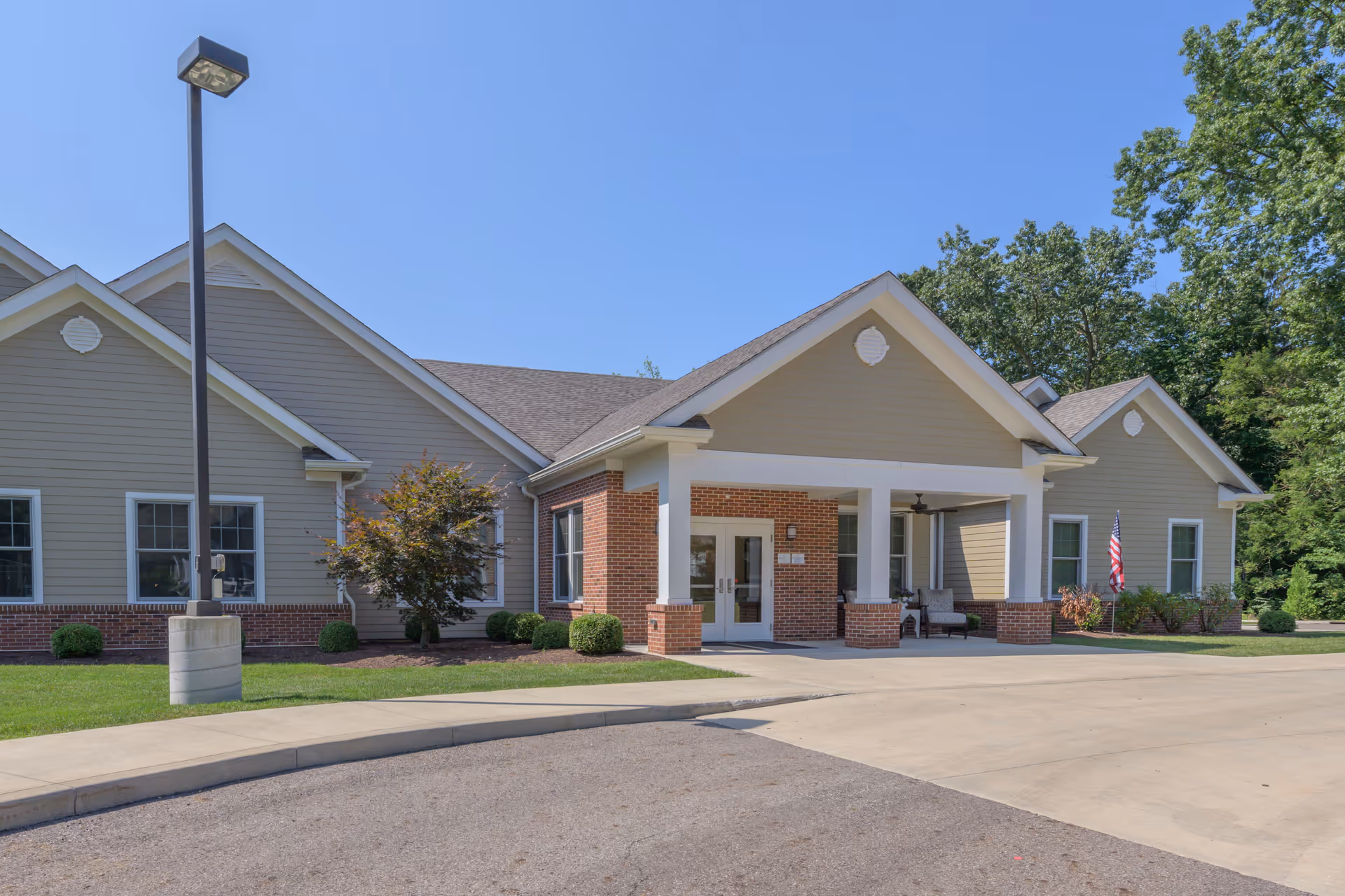 Exterior view of a single-story senior living facility building with beige siding and brick accents, a covered entrance with white columns, a small porch area with seating, a flagpole with an American flag, and a clear blue sky above.