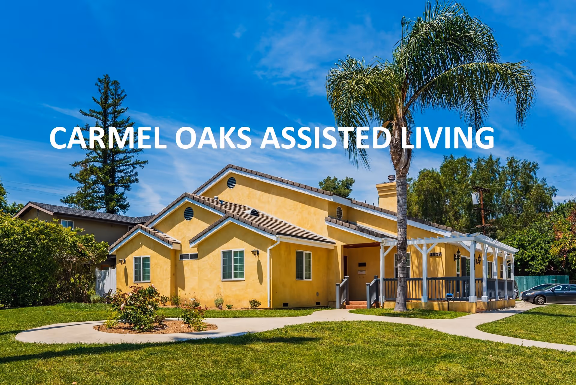 Exterior view of Carmel Oaks Assisted Living facility showing a yellow building with a tiled roof, a covered porch, a palm tree, green lawn, and a clear blue sky.