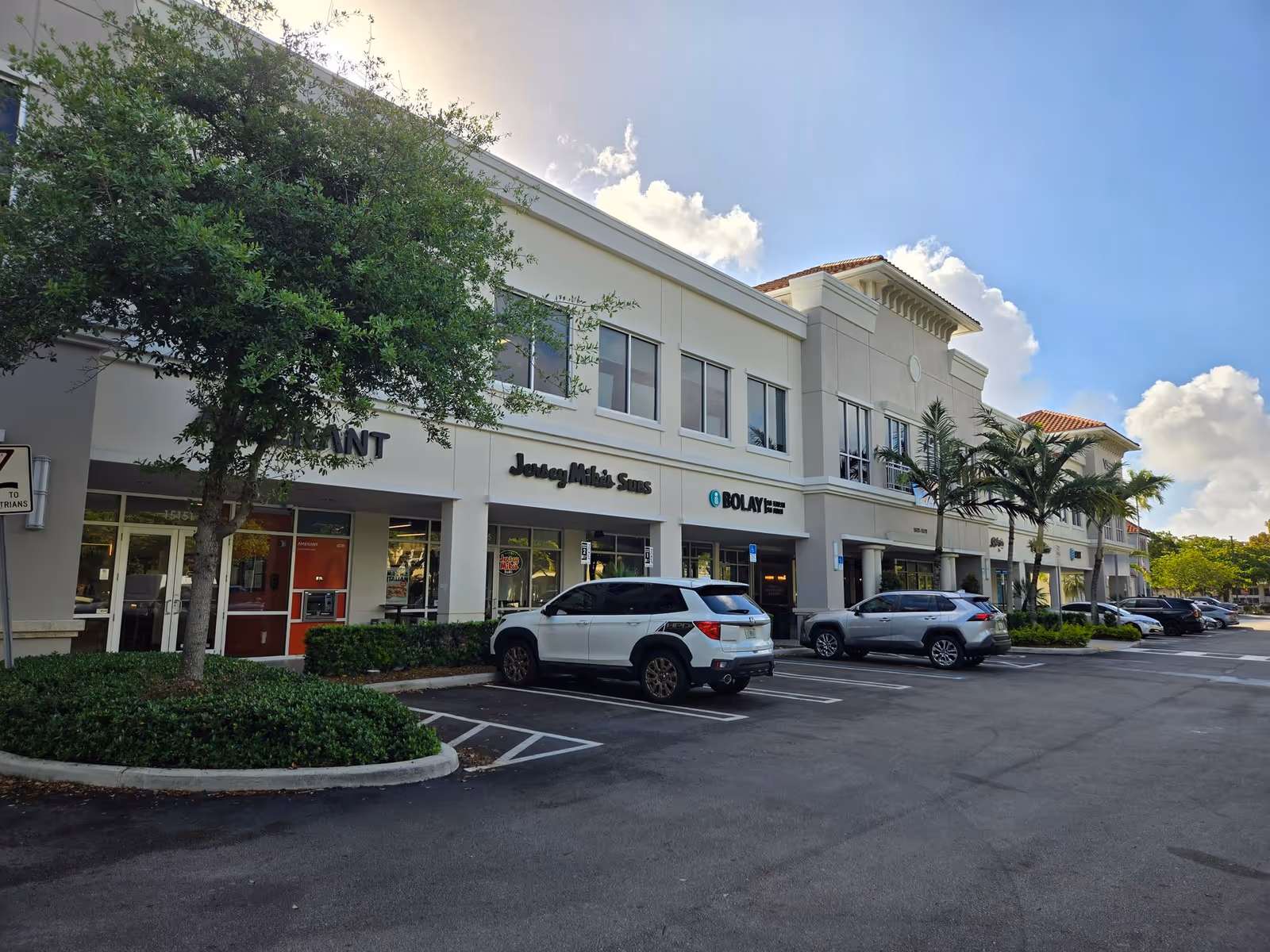 Shopping plaza exterior with storefronts, parked cars, and palm trees under a blue sky.