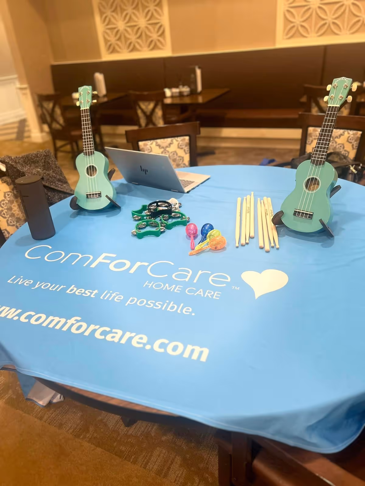 Table setup featuring teal ukuleles, laptop, and promotional ComForCare items in an interior setting.