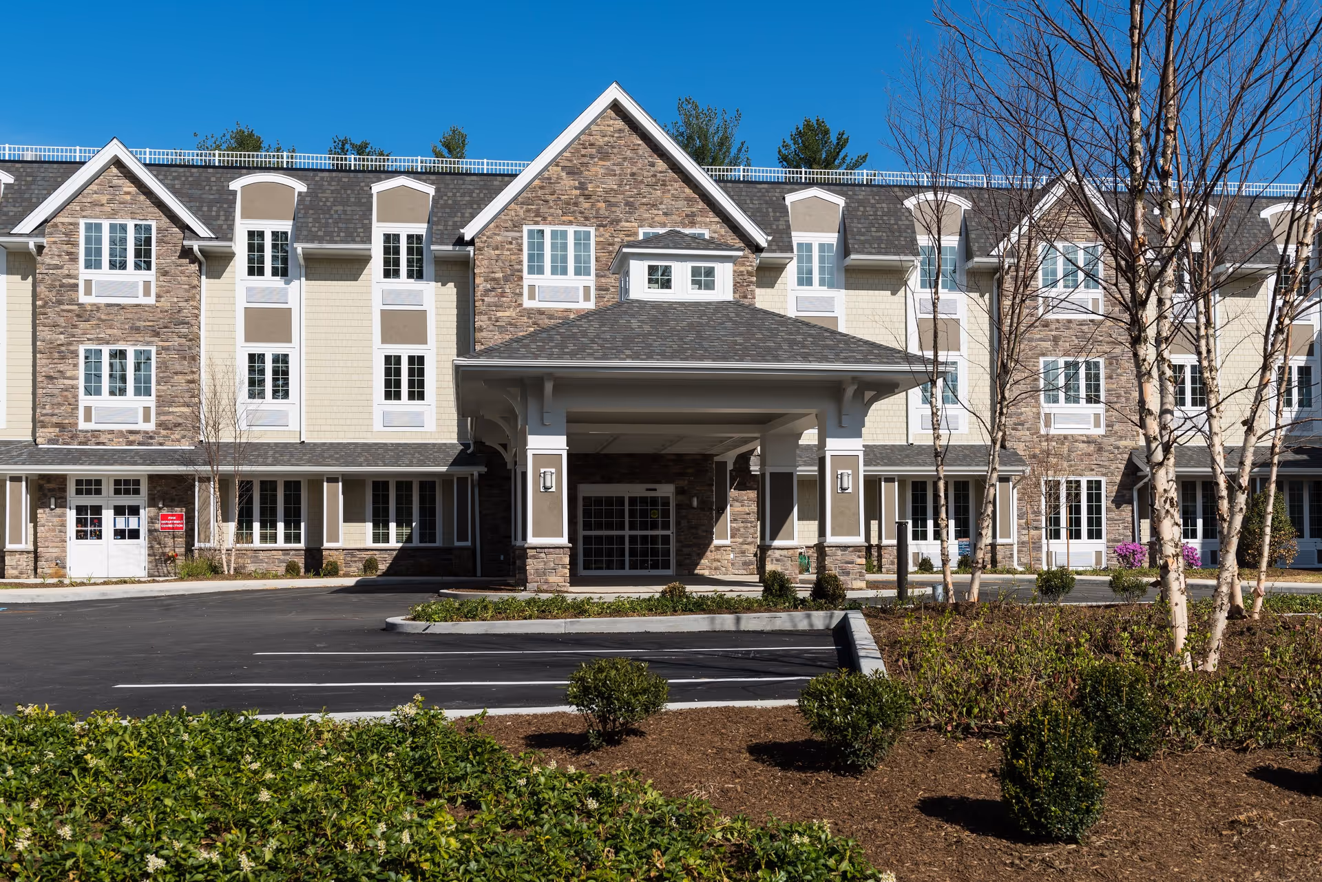 Front exterior of a three-story senior living facility with a covered entrance, parking lot, and landscaped grounds.