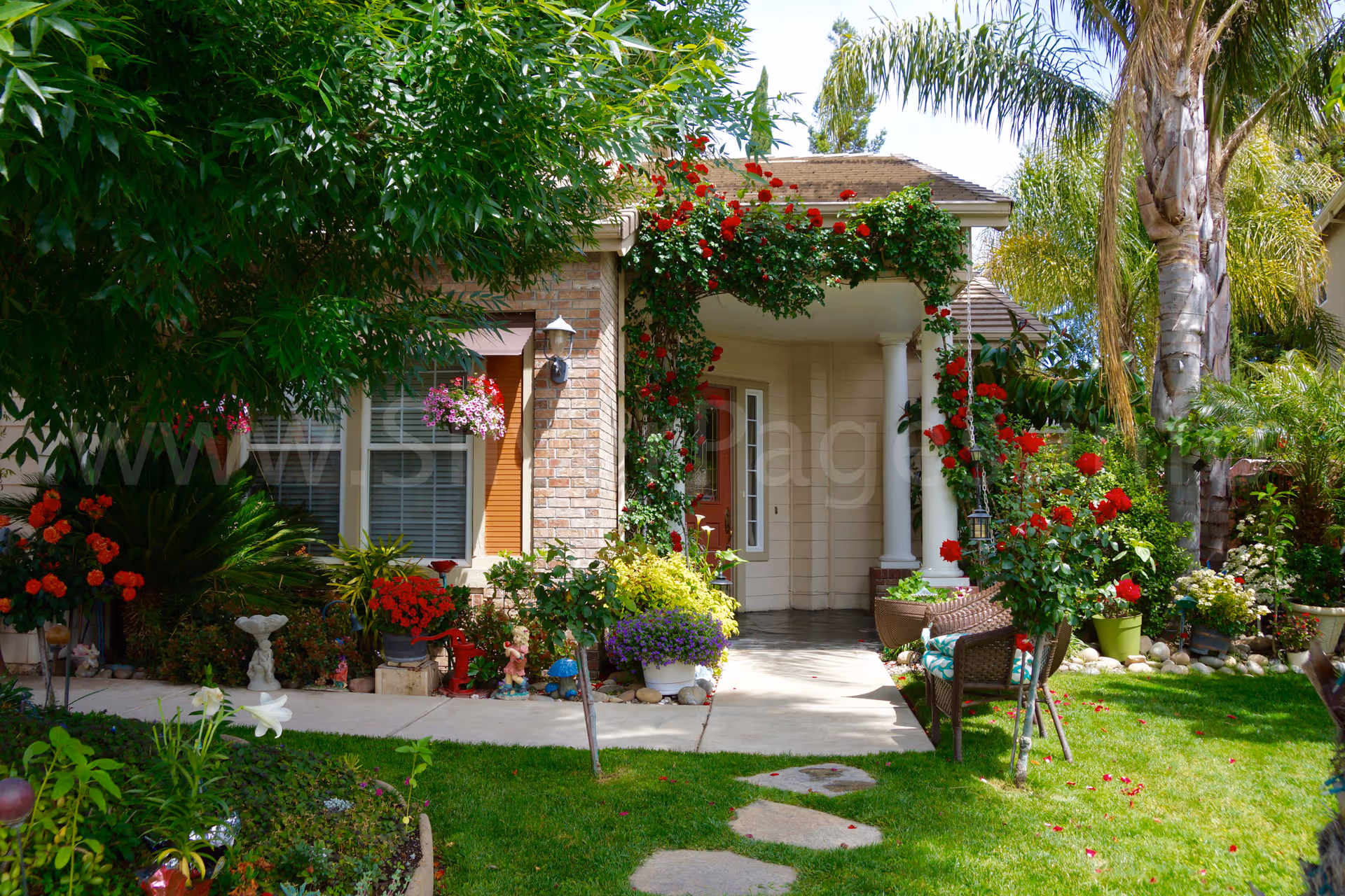A sunny residential entrance with a flowering garden and seating on a welcoming front porch.