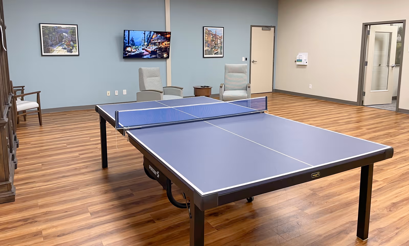 Blue ping-pong table in a hardwood-floored recreational room with chairs and framed wall art.