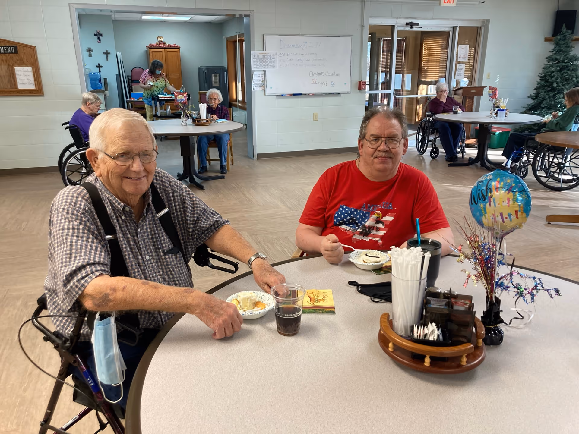 Two older men seated at a round table eating dessert in a senior living communal dining room, with other residents at tables in the background.