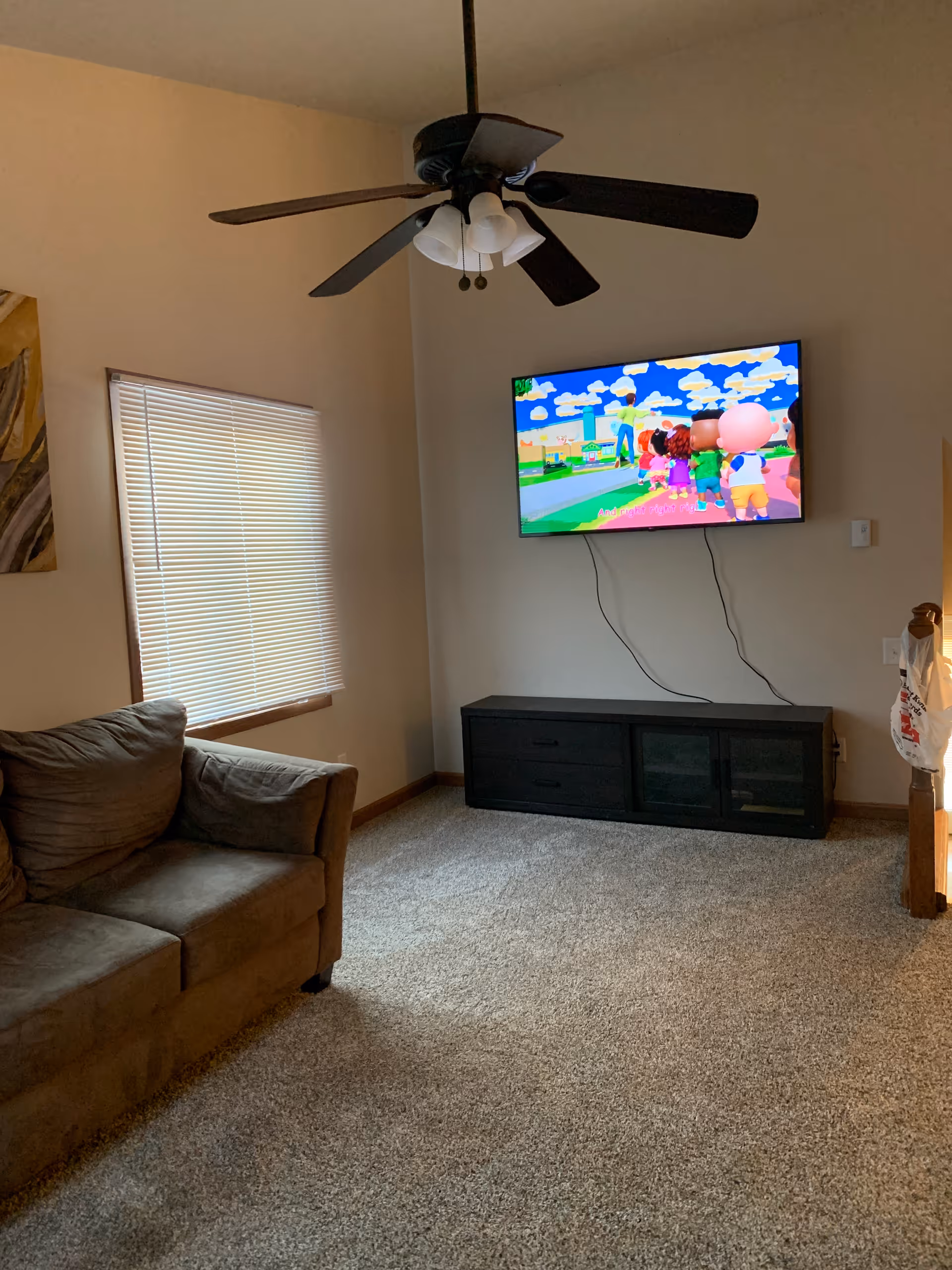 Living room with sofa, ceiling fan, blinds, and mounted TV.