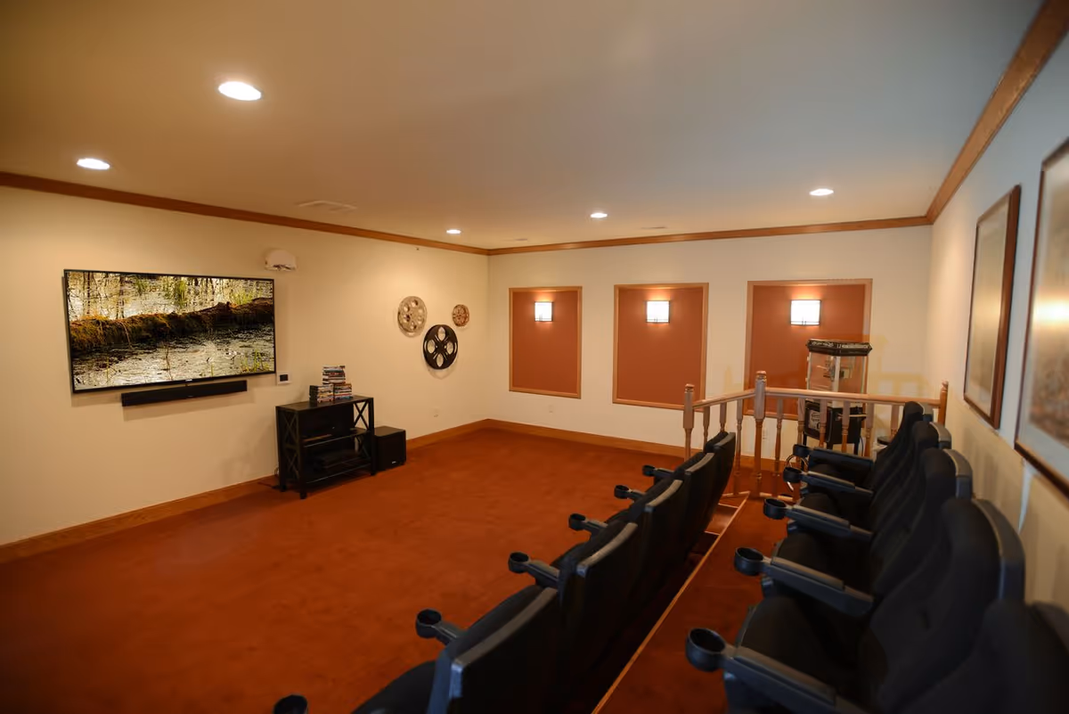 A small home theater room with a row of black cushioned seats facing a wall-mounted flat-screen TV. The room has warm lighting with recessed ceiling lights, brown carpet, and beige walls with wooden trim. There is a popcorn machine in the corner and decorative film reels on the wall.
