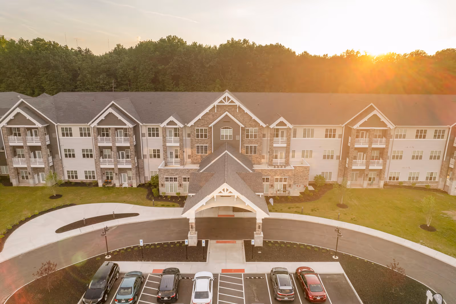 Front exterior of a multi-story senior living building with a covered porte-cochère entrance, parking lot and cars at sunset.