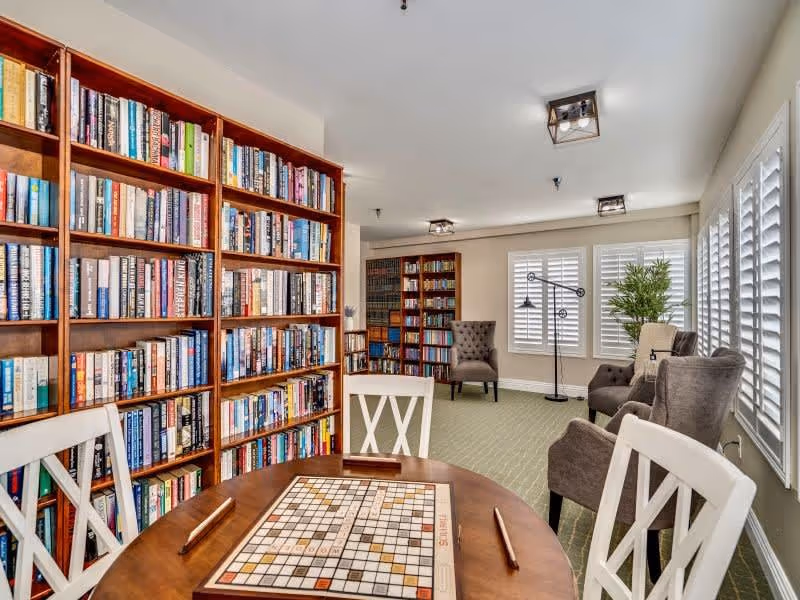 A cozy reading and game room with large wooden bookshelves filled with books, a round table with a Scrabble board and four chairs, comfortable armchairs, large windows with white shutters, and a potted plant in the corner.