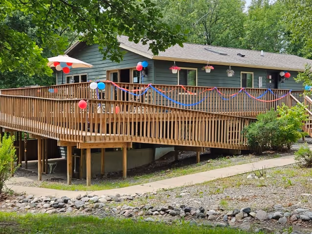 A single-story building with a large wooden deck decorated with red, white, and blue balloons and streamers. The building is surrounded by trees and greenery, with a stone-lined pathway leading up to it. Hanging flower pots are visible on the deck railing.