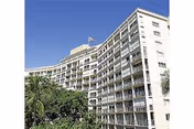 A curved, multi-story retirement residence with balconies, palm trees, and a clear blue sky.