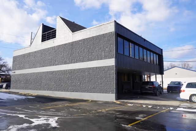 Exterior of a two-story building with dark brick facade and large windows, parking area.