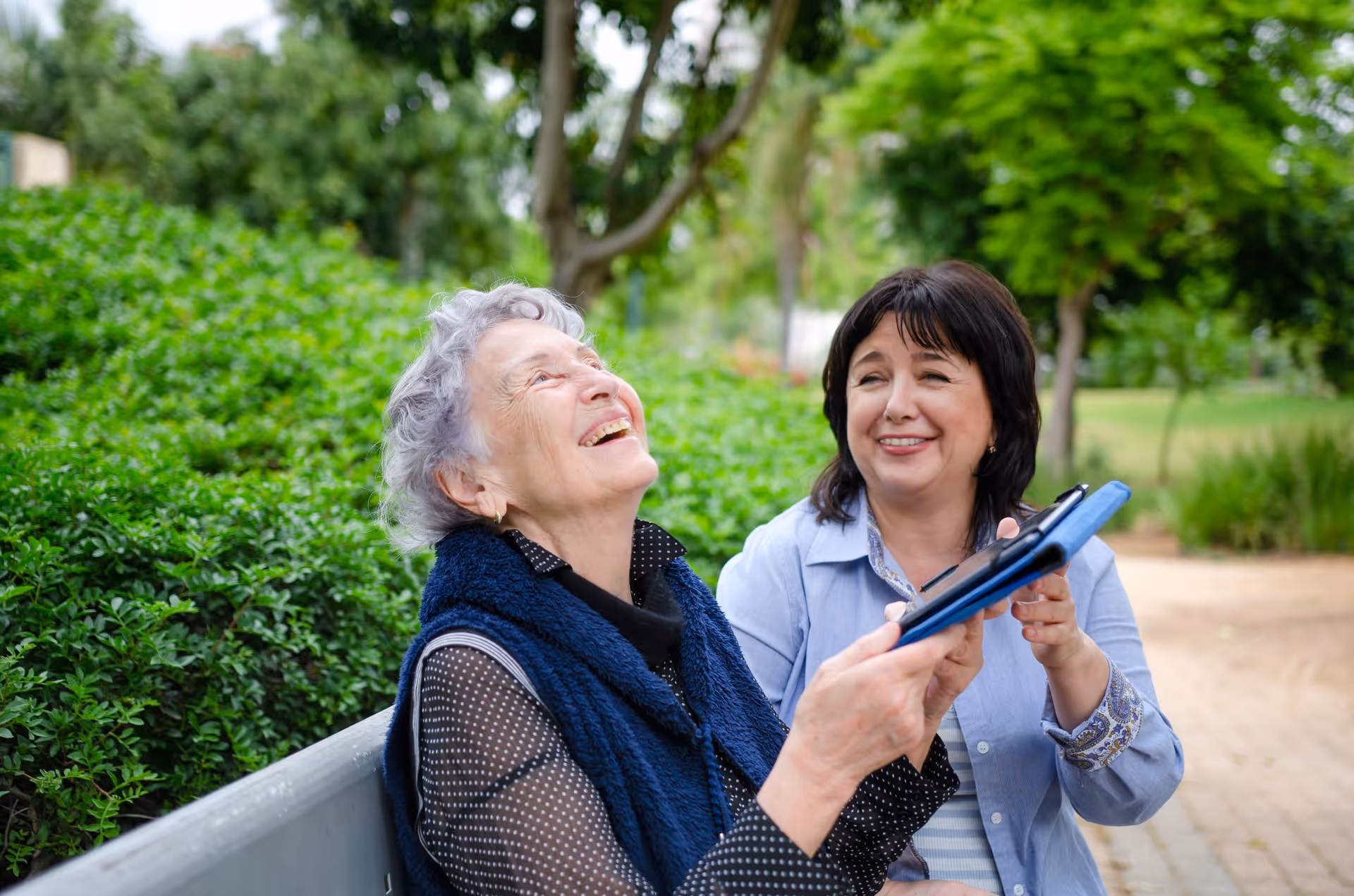 Two smiling women sit on an outdoor bench, one showing a tablet to the other.