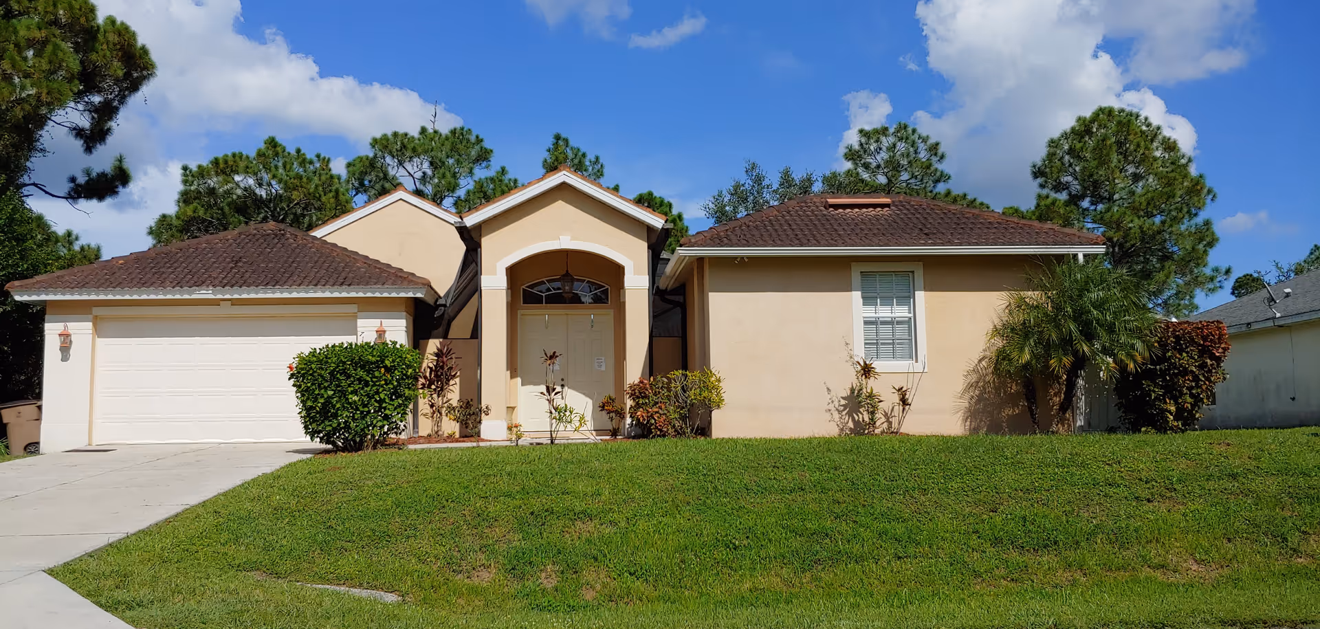 Single-story beige house with an attached garage, front lawn and palm trees under a blue sky.