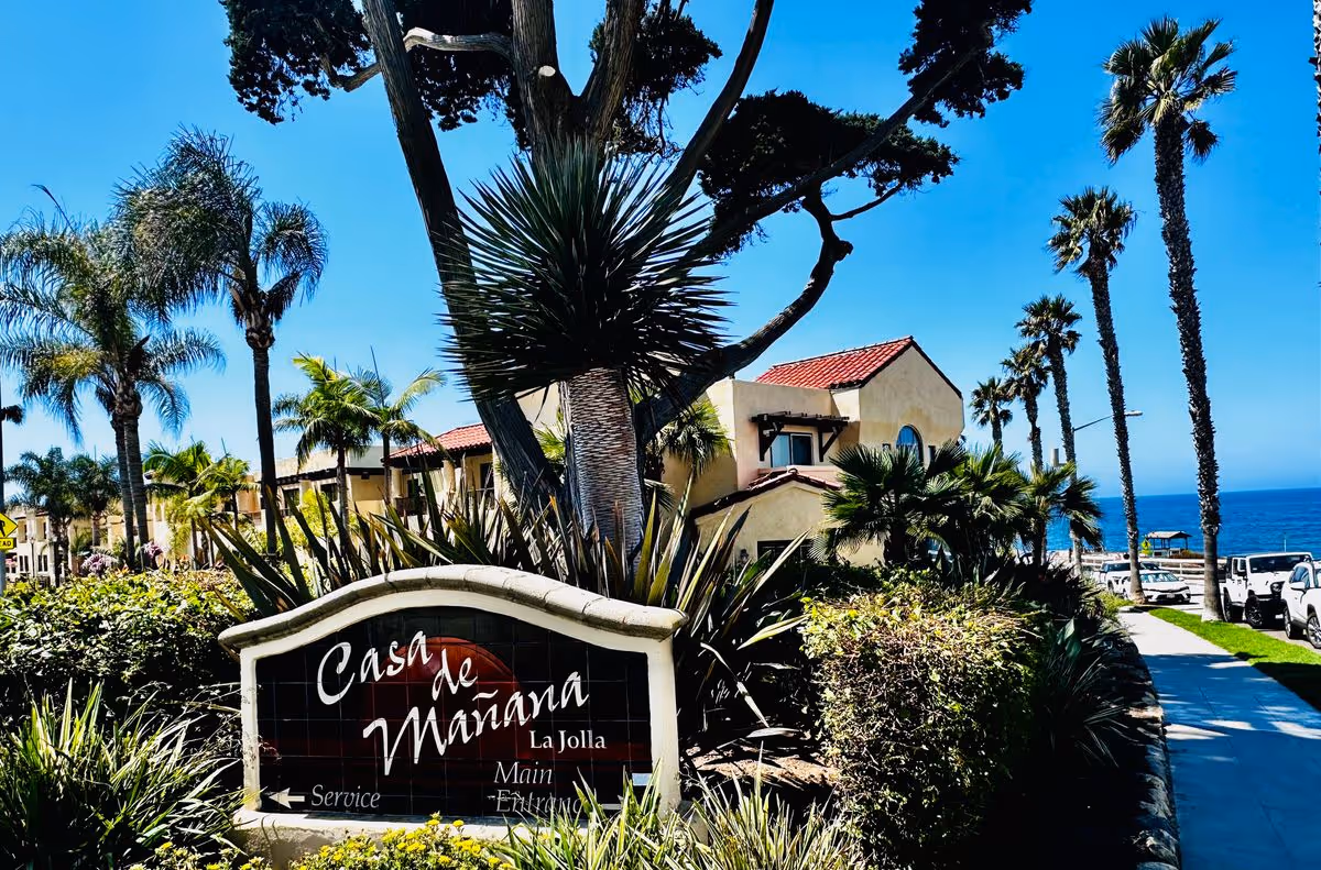 Entrance sign reading "Casa de Mañana" in front of palm trees and Mediterranean-style buildings with the ocean in the background.