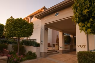 Sunlit exterior entrance of San Carlos Elms with pillars, greenery, and a bench.