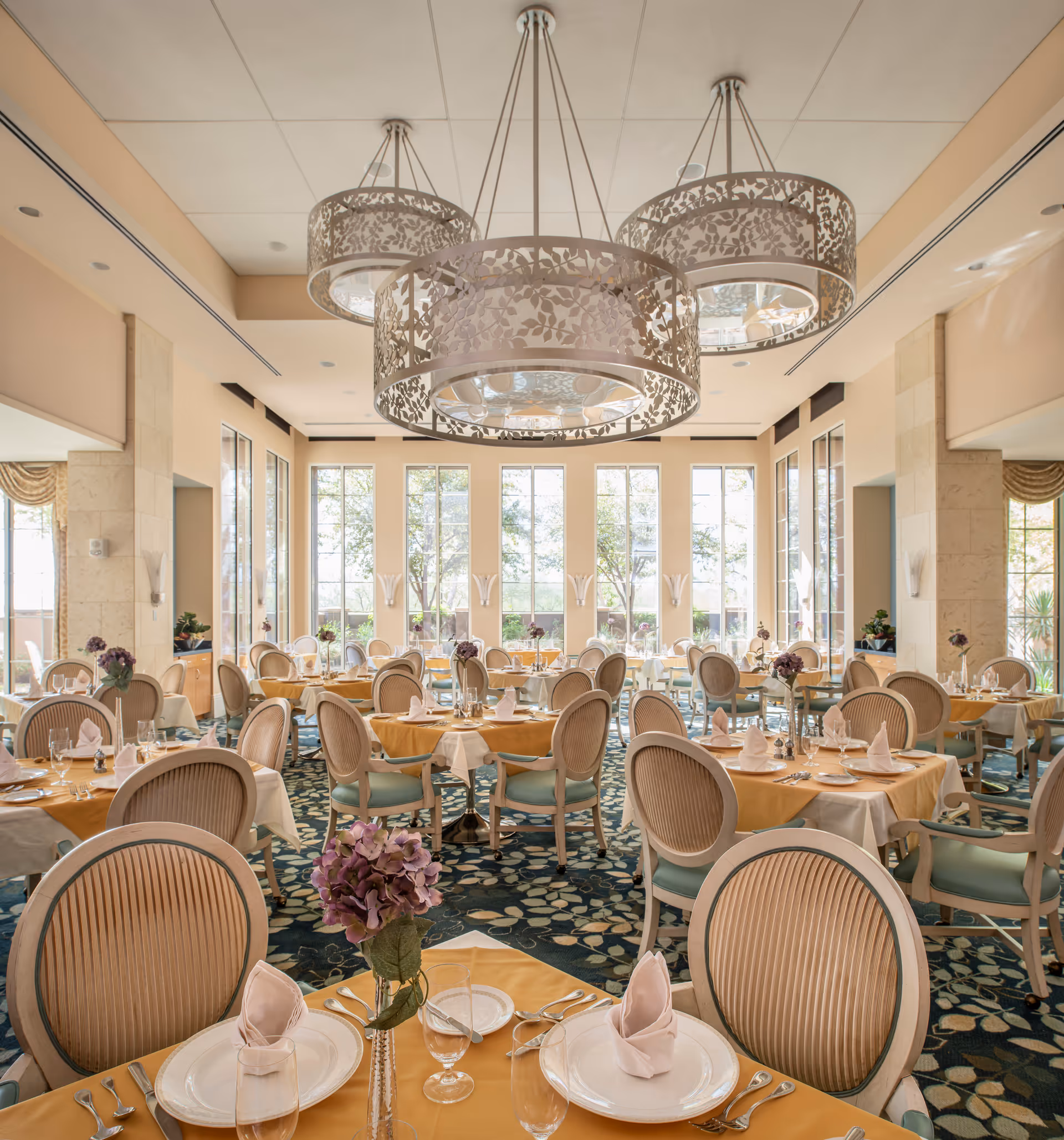 Elegant dining room with round tables set with plates and folded napkins, upholstered chairs, large decorative chandeliers, and tall windows letting in natural light.