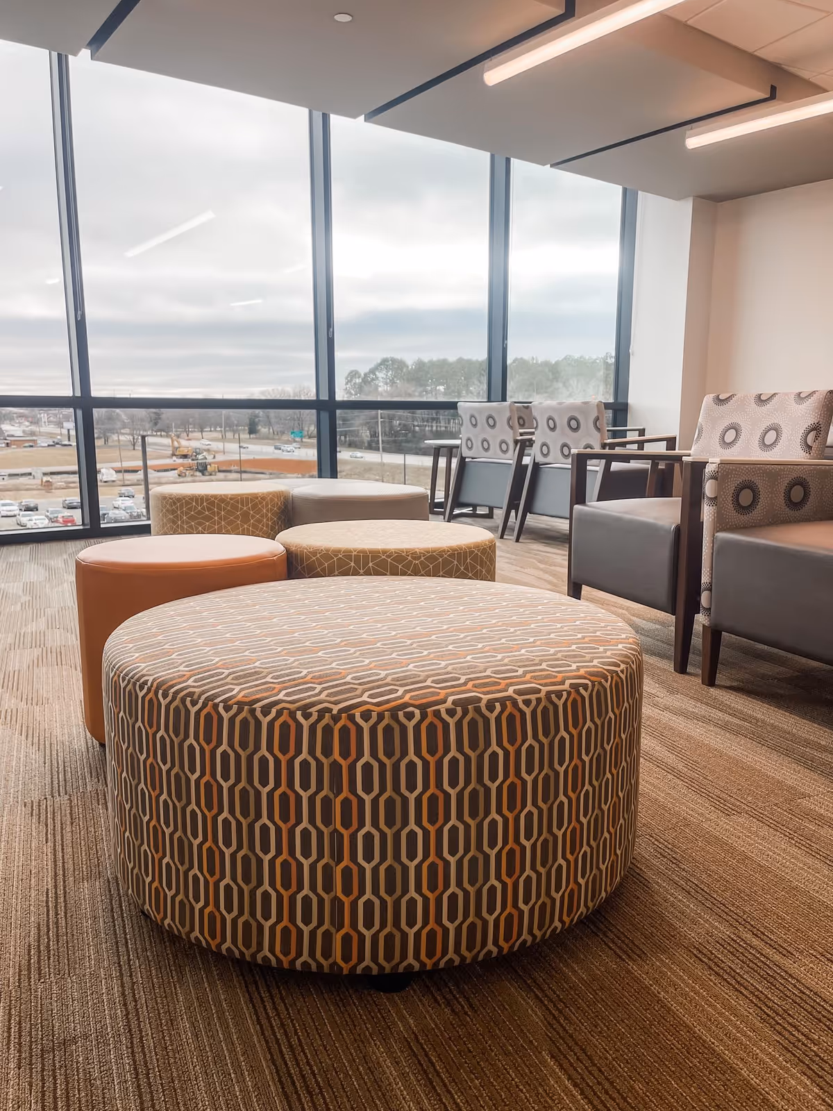 Bright, modern lounge area with patterned ottomans and armchairs beside large windows.