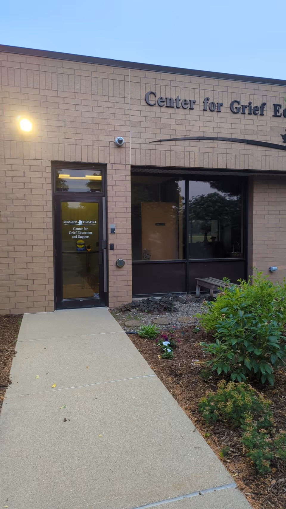 Brick exterior of Seasons Hospice entrance with glass doors and landscaped walkway.