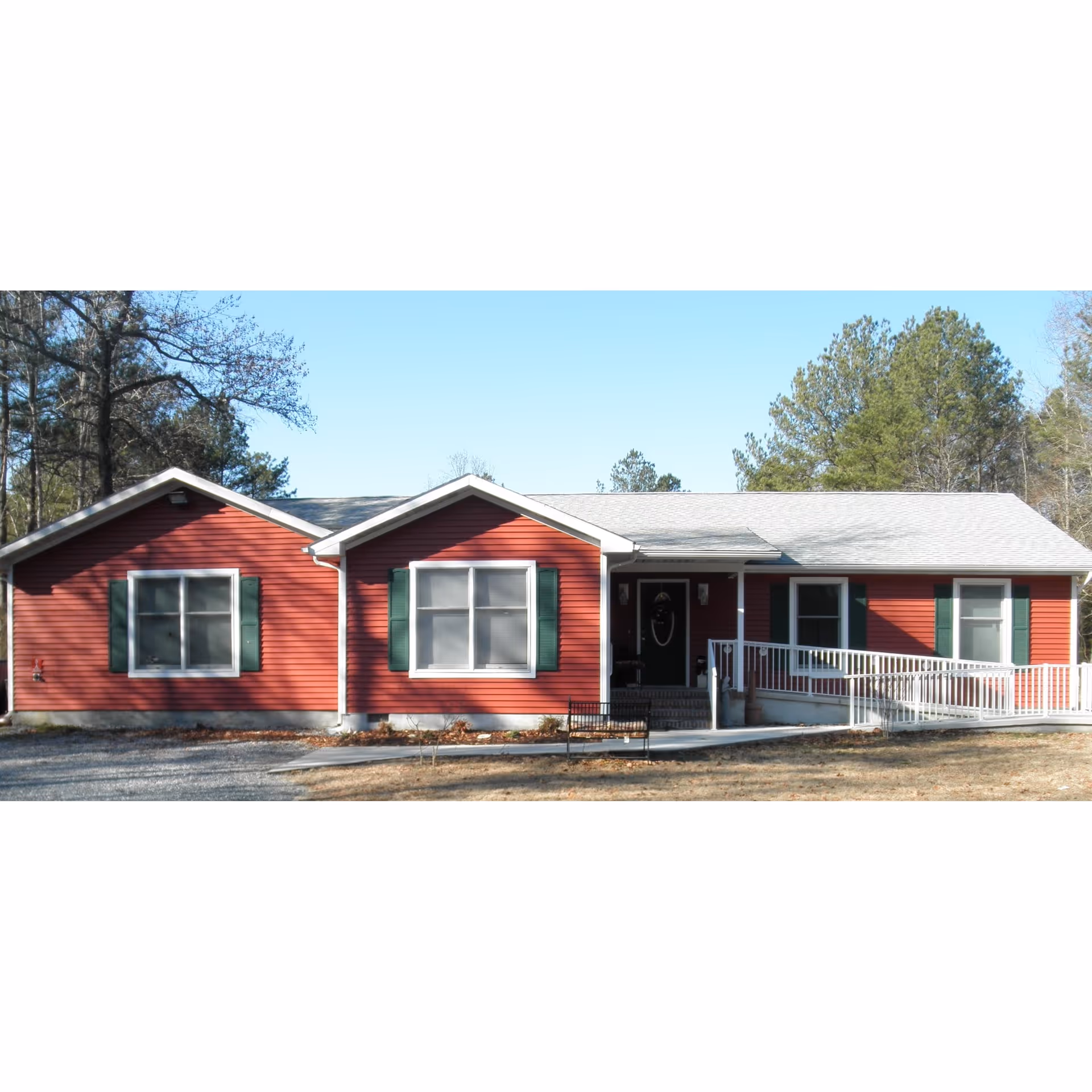 Front view of a single-story red building with white trim, green shutters, a front porch and wheelchair ramp surrounded by trees.