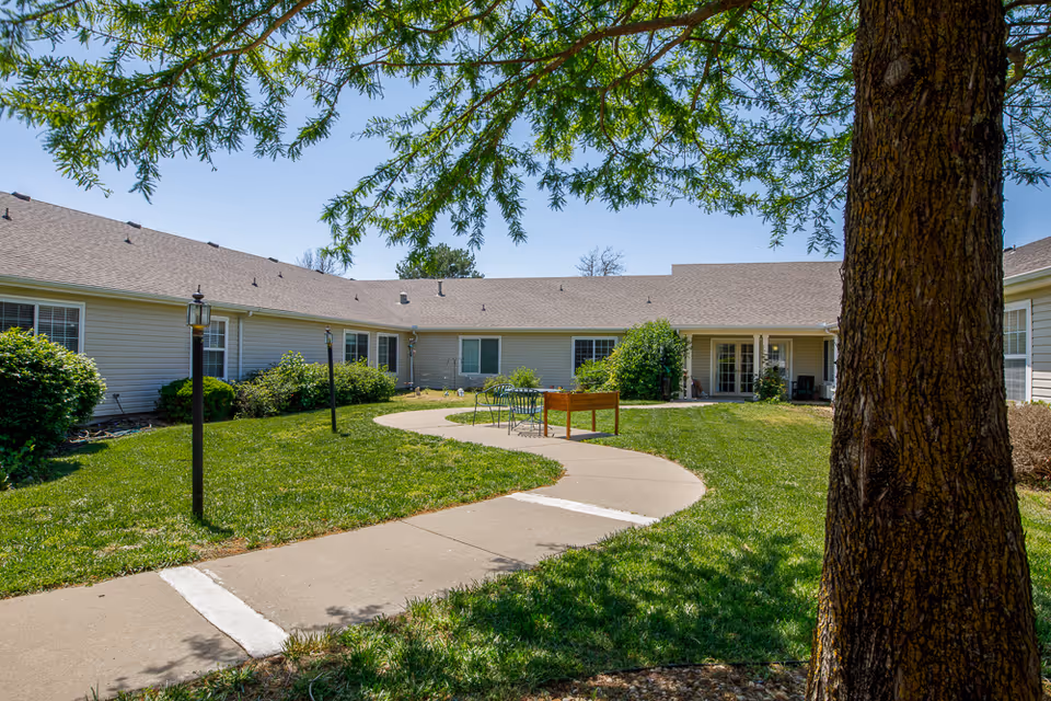 Outdoor courtyard area at Homestead Assisted Living of Abilene with a curved concrete walkway, green grass, bushes, and a large tree in the foreground. The single-story building with light-colored siding surrounds the courtyard, and there are outdoor chairs and a table near the center.