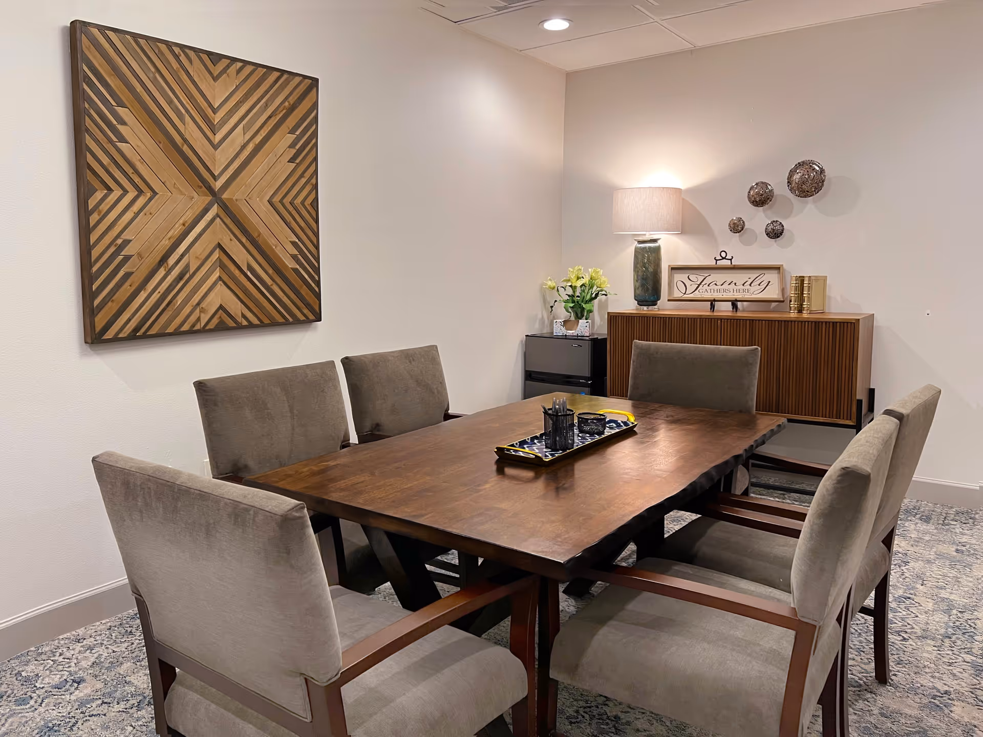 A dining room with a wooden table surrounded by six upholstered chairs. On the table is a decorative tray with some items. Against the wall is a wooden sideboard with a lamp, a vase with flowers, decorative wall art, and a sign that reads 'Family Gathers Here'. A large geometric wooden wall art piece hangs on the adjacent wall. The room has a patterned carpet and soft lighting.