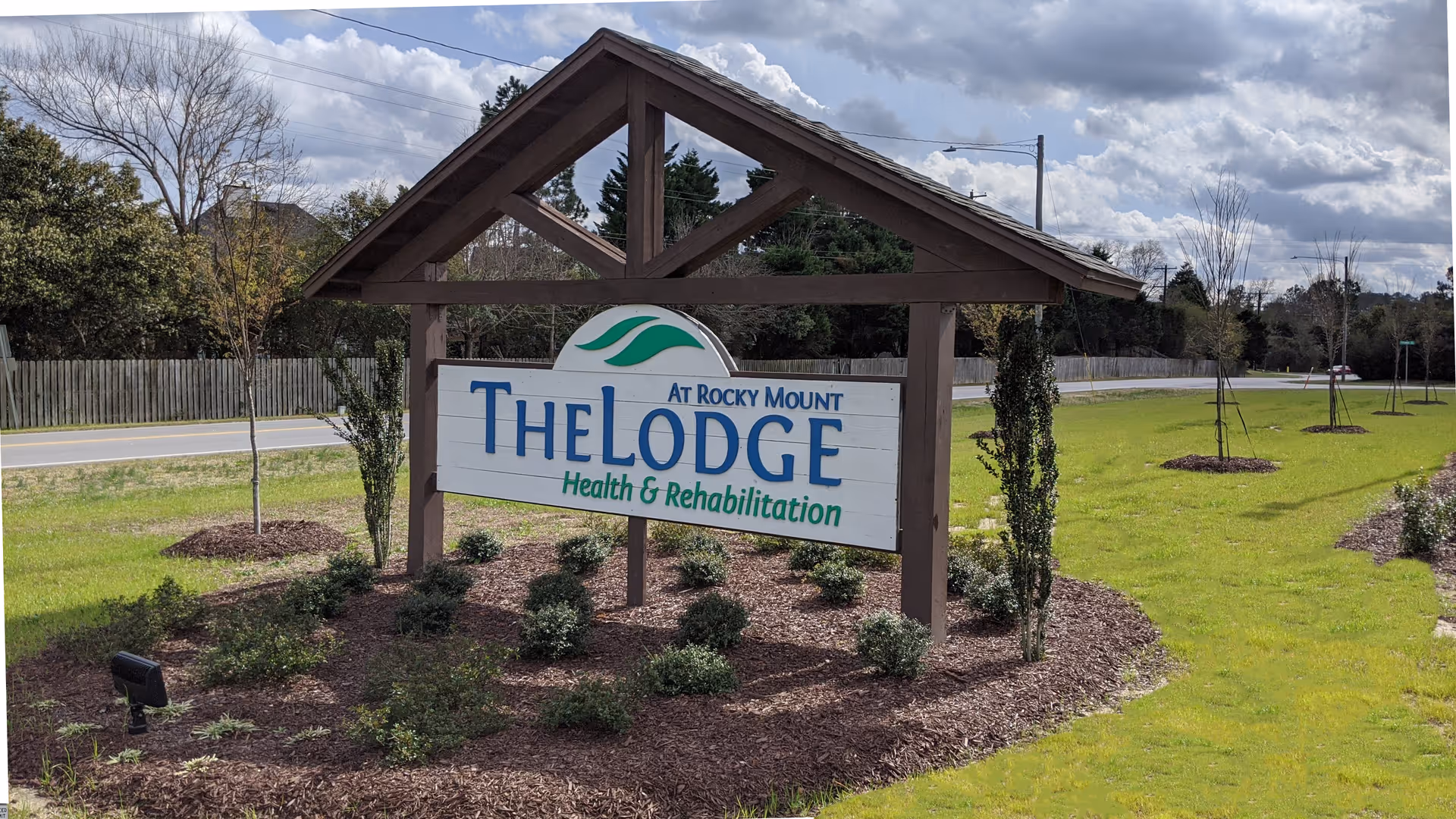 Outdoor view of a wooden sign for The Lodge at Rocky Mount Health & Rehabilitation, surrounded by landscaped bushes and grass, with a road and trees in the background under a partly cloudy sky.