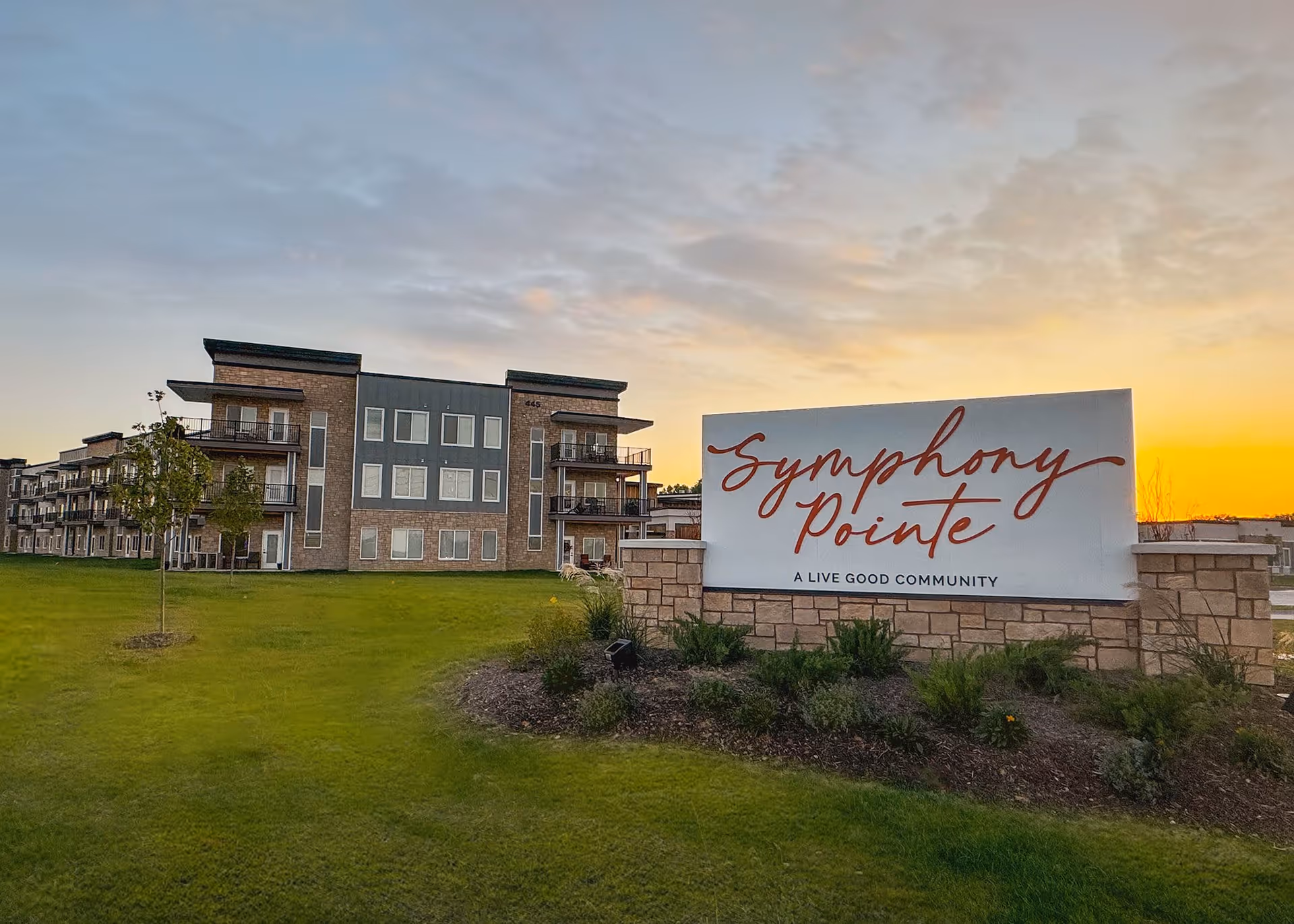 The Symphony Pointe sign in front of a multi-story senior living building on a grassy lawn at sunset.