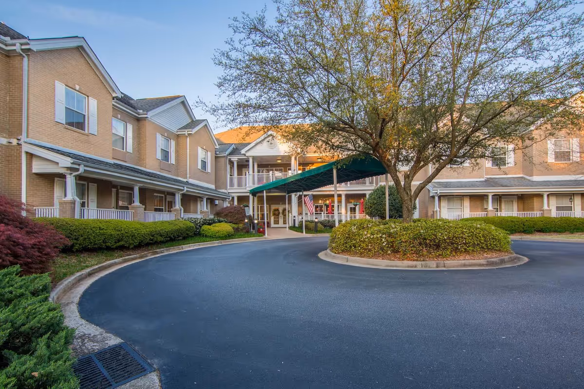 Exterior view of The Gardens at Eastside senior living facility showing a two-story brick building with white trim, a covered entrance with a green canopy, an American flag, and landscaped bushes and trees around a circular driveway.